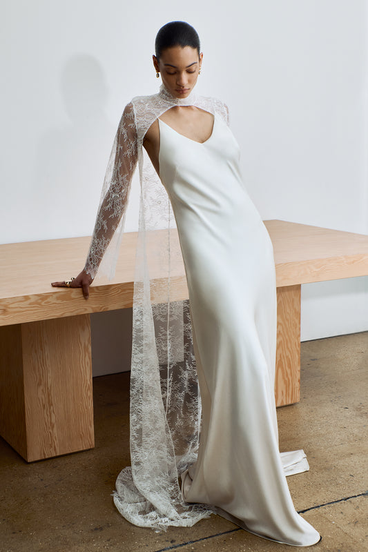 Woman wearing a white wedding dress with a sheer long sleeve lace cape in a minimalistic setting.