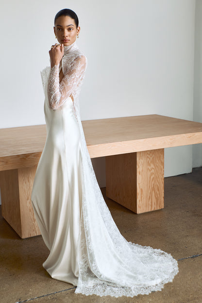 Woman wearing a white wedding dress with a long sleeve lace cape overlay in a minimalistic setting.